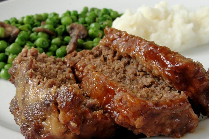 three slices of Chef John's glazed meatloaf on a plate with mashed potatoes and peas with mushrooms in the background
