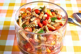 a colorful pasta salad in a glass bowl sitting on an orange and yellow gingham tablecloth