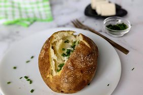 close up view of a baked potato, garnished with with chives on a white plate, with a fork, butter and chives in the background 