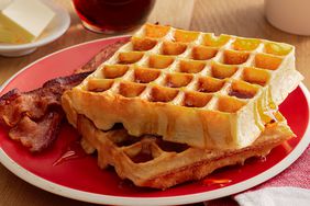 a low angle, close up view of a two square waffles stacked on a red plate, topped with syrup, and served with bacon.