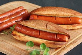 close up view of two Basic Air Fryer Hot Dogs in toasted buns, next to three hot dog sausages, on a wooden cutting board garnished with fresh herbs 