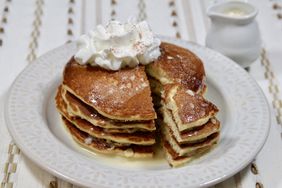 golden stack of four pancakes with a wedge-shaped bite cut out, topped with whipped cream on white plate and gold and white tablecloth