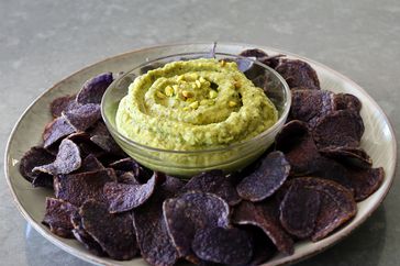 Green zucchini hummus garnished with pistachios in glass bowl surrounded by blue potato chips
