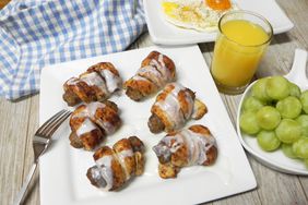 sausages baked in cinnamon roll wrapping with icing on white plate with full breakfast in background