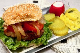 beautifully grilled turkey burger with lettuce, tomato, and grilled onions on bun on plate with pickles, chips, banana peppers, and radish