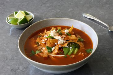 white bowl of chicken tortilla soup with deep red broth, tortilla strips, avocado slices, and crumbled cheese