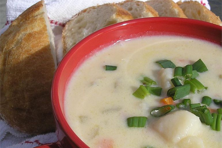 a red bowl of creamy soup with chunks of potato, garnished with sliced green onions with sliced bread in the background