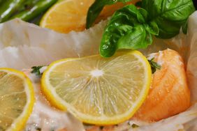 close up view of a salmon fillet with a lemon slice and herbs on parchment paper, with fresh basil leaves