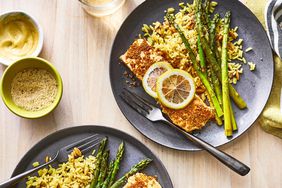 Breaded, baked salmon fillets topped with lemon slices, served alongside asparagus slices and rice pilaf on blue plates
