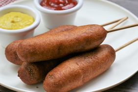 four corn dogs on a white plate with ramekins of mustard and ketchup in the background